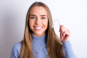 Young Caucasian girl wearing blue turtleneck over white background holding an invisible braces aligner recommending this new treatment.